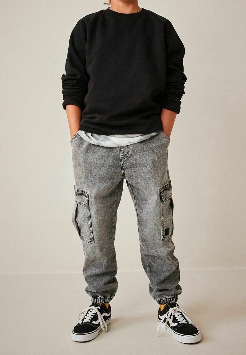 Personne vêtue d’un sweat-shirt noir, d’un jean cargo gris avec des poignets élastiques et de baskets noires et blanches, debout les mains dans les poches.