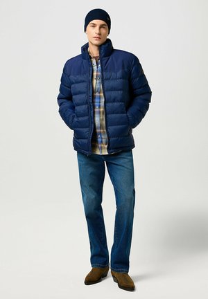 Doudoune marine avec col haut, texture matelassée, portée sur une chemise en flanelle à carreaux multicolores, associée à un jean bleu et des bottes marron.
