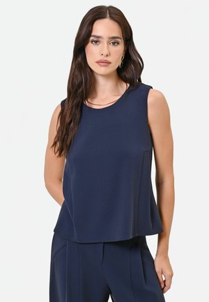 Donna con lunghi capelli scuri che indossa una canotta blu navy e pantaloni abbinati, in piedi davanti a uno sfondo chiaro con un'espressione neutra.