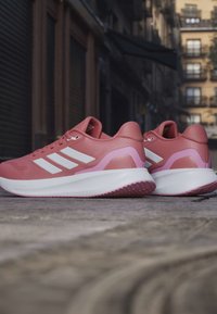 Paire de chaussures de sport roses et blanches avec des bandes blanches, posées sur un pavé en pierre dans un environnement urbain.