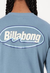 Blå bomullssweatshirt med en stor hvit "Billabong"-logo med blå kontur og et stilisert bølgedesign under.
