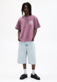 Fialové oversized tričko s bielou grafikou na hrudi, skombinované s svetlomodrými denimovými šortkami. Čierne topánky a biele ponožky dotvárajú outfit.