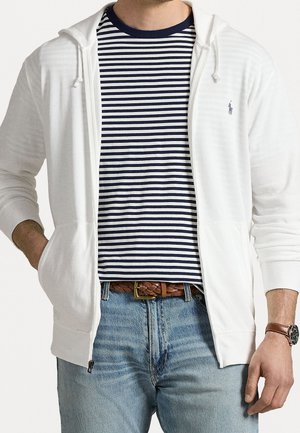 Man wearing a white zip-up hoodie over a navy and white striped shirt, light blue jeans, brown braided belt, and brown leather watch.