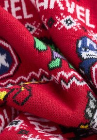Roter gestrickter Stoff mit weißen und schwarzen Mustern, zudem bunten geometrischen Formen in Grün, Lila und Gelb, die strukturierte Details hervorheben.