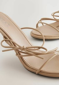 Beige High Heelsandalen verfügen über dünne, verwobene Riemen, eine glatte Fußbett und ein rundes Zehendesign mit einem einfachen Knotendetail vorne.