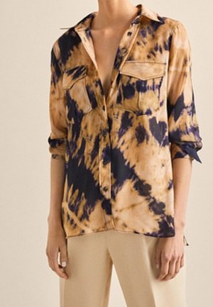 Frau trägt ein lockeres Hemd mit Knöpfen, das ein abstraktes Batikmuster in Schwarz und Beige aufweist, und beige Hosen, die Ärmel sind bis zu den Ellbogen hochgekrempelt.