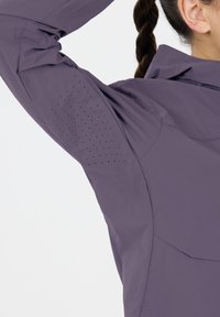 Personne portant une veste violette avec des trous de ventilation sous le bras levé, montrant des cheveux tressés et une oreille sur fond blanc.