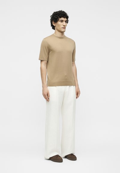 Νέος άνδρας που στέκεται κοιτάζοντας μπροστά, φοράει μπεζ κοντομάνικο πουκάμισο, λευκό παντελόνι με φαρδιά πόδια και καφέ παπούτσια σε μονόχρωμο φόντο.