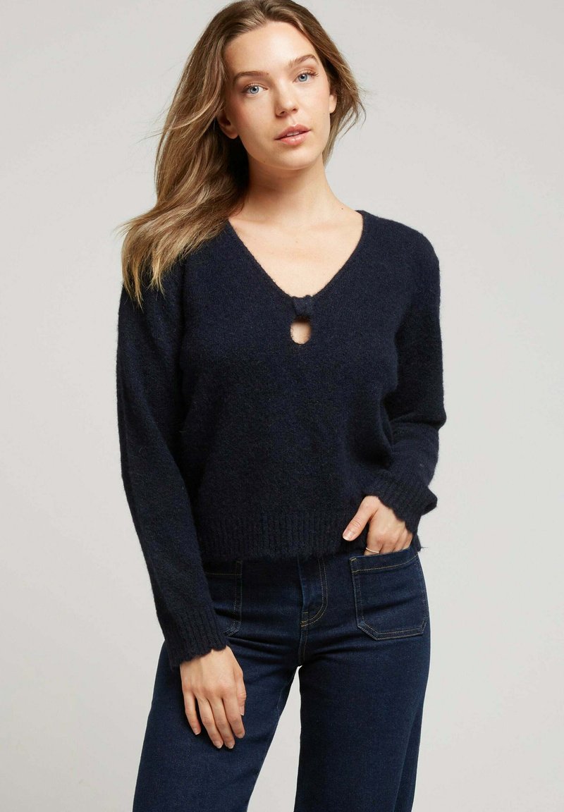 Pull en tricot bleu marine avec une coupe ample, col en V et un détail de trou central. Texture douce, ourlet côtelé et manches longues. Associé à un jean foncé.