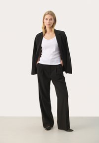 Černý strukturovaný blazer přes bílý top bez rukávů, v kombinaci s volnými černými kalhotami a černými botami s špičatou špičkou. Hladká tkanina a čisté linie.
