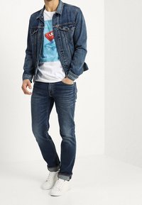 Jeansowa kurtka na białym T-shircie z grafiką przedstawiającą niebieski i czerwony wzór, w połączeniu z ciemnymi dżinsami i białymi trampkami.