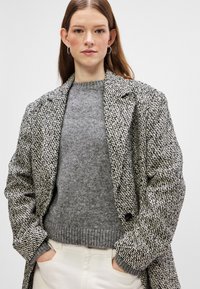 Villasegu villasegowool segmendi kohtu mustvalge houndstooth mustri, lõdva lõike, eenduvate kraede ja nööbiga kinnitusega. Hall ümarkaelusega sviiter all.