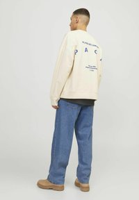 Moški oblečen v bež pulover s modrim napisom, ohlapne modre kavbojke in svetlo rjave čevlje, stoji obrnjen stran od kamere ob enobarvni svetli ozadju.