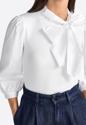 Femme portant un chemisier blanc avec des manches trois-quarts bouffantes et un grand nœud au col, associé à un pantalon en denim bleu foncé taille haute.