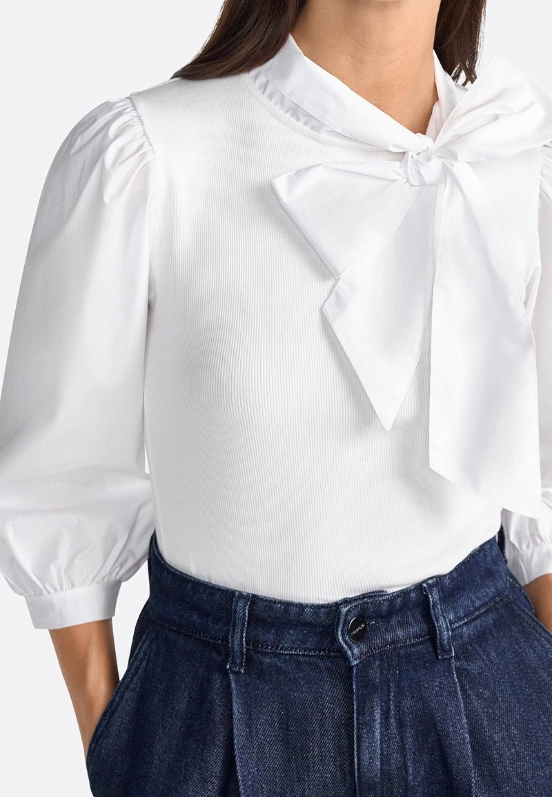Femme portant un chemisier blanc avec des manches trois-quarts bouffantes et un grand nœud au col, associé à un pantalon en denim bleu foncé taille haute.