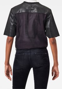 Bakifrån av en person som bär en svart kortärmad topp med läderaxlar och transparent randigt tyg, tillsammans med mörka smala jeans.