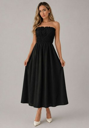 Femme portant une robe maxi noire bustier froncée, tenant la jupe, associée à des talons blancs pointus sur un fond gris.