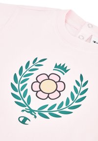 Camiseta de color rosa claro con un diseño central de una flor estilizada con un centro amarillo y pétalos rosas, rodeada de hojas verdes y una corona.