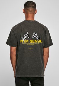 Tamsiai pilkas medvilnės marškinėliai su grafika, vaizduojančia dvi šachmatų vėliavas, ir tekstu geltona spalva. Trumpi rankovės ir laisvas kirptis.