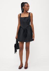 Melnas satīna kleitas ar kvadrātveida izgriezumu, pieguļošu augšdaļu un savāktu svārku. Modelis tur mazu melnu somu un valkā bordo krāsas papēžus.