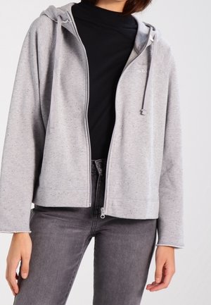 Personne portant un sweat à capuche zippé gris clair sur un t-shirt noir et un jean gris foncé, debout, les bras détendus le long du corps.