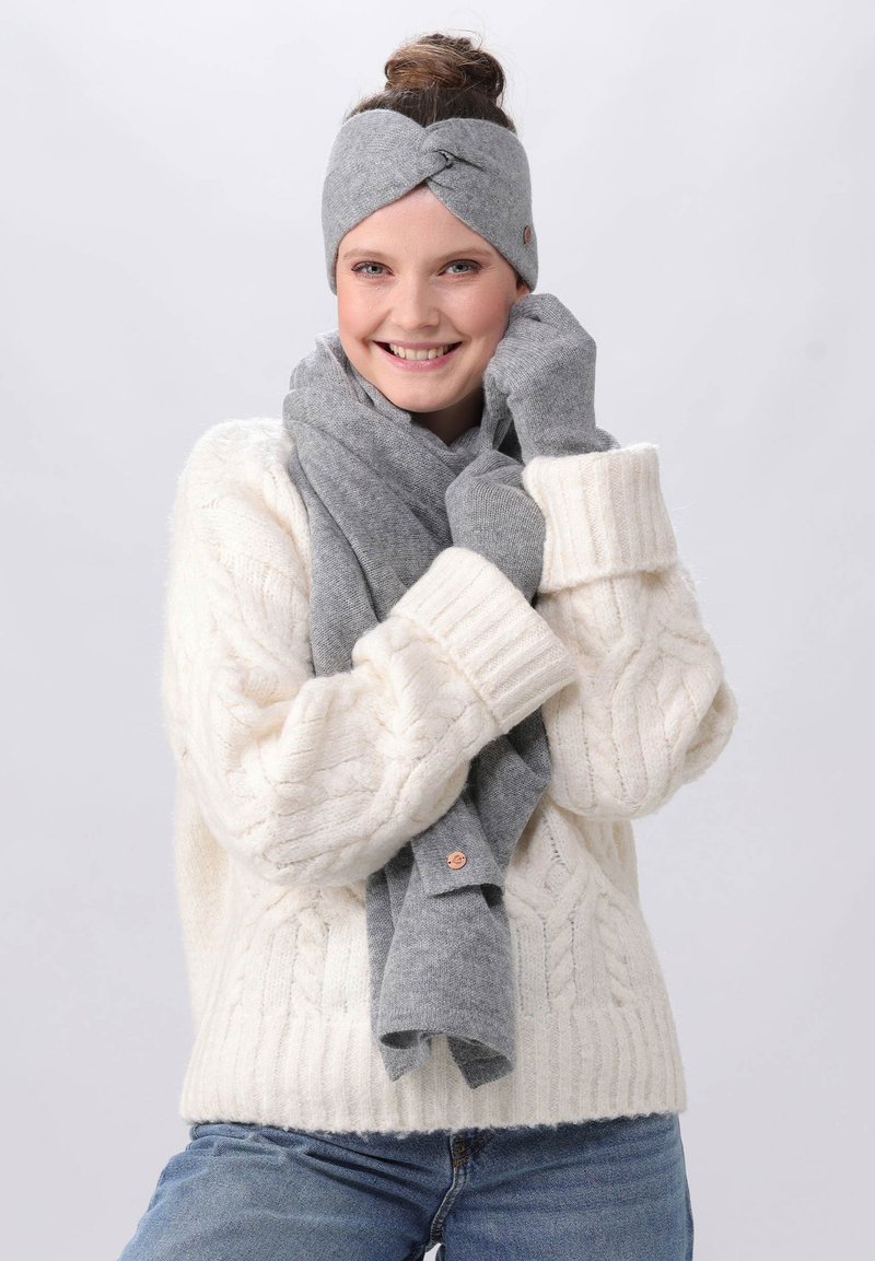 Un bandeau en tricot gris avec un design torsadé, un écharpe grise assortie et des gants sans doigts, portés avec un pull crème en tricot torsadé.