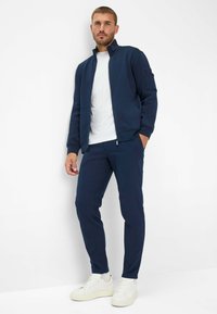 Marinefarbener Zip-Jacke mit hohem Kragen, kombiniert mit passenden marinefarbenen Hosen und weißen Sneakern. Glattes Material und taillierte Passform.