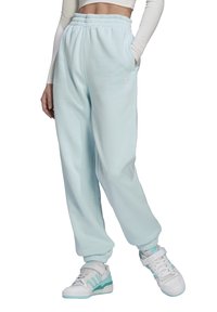 Osoba w jasnoniebieskich dresowych spodniach Adidas z elastycznym pasem i mankietami, w połączeniu z białymi i turkusowymi butami Adidas, z rękami w kieszeniach.
