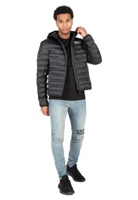 Schwarze Pufferjacke mit Kapuze, mit Reißverschluss und gestepptem Muster. Kombiniert mit hellblauen, abgetragenen Jeans und schwarzen Sneakers.