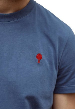 Primo piano di una persona che indossa una maglietta blu con un piccolo albero rosso ricamato sulla parte sinistra del petto.