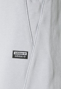 Svetle šedá mikina s texturovanou tkaninou. Obsahuje černou nášivku loga Adidas se dvěma horizontálními liniemi a třemi listovými emblémy.