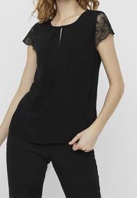 Mujer vistiendo una blusa negra con mangas de encaje y un pequeño escote en forma de llave, combinada con pantalones negros frente a un fondo liso.