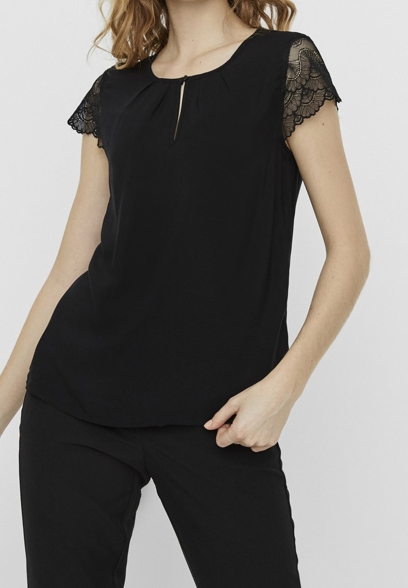 Mujer vistiendo una blusa negra con mangas de encaje y un pequeño escote en forma de llave, combinada con pantalones negros frente a un fondo liso.