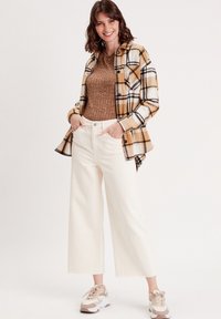 Camisa de cuadros en amarillo, negro y blanco sobre un top marrón de canalé; combinada con pantalones anchos blancos y zapatillas beige con paneles texturizados.
