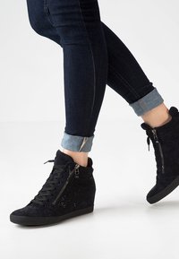 Baskets compensées noires en suède avec des accents scintillants, dotées d'une fermeture éclair et de lacets. Portées avec un jean en denim foncé retroussé.