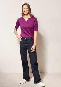 Krátký rukáv fialového trička s výstřihem do V, v kombinaci s tmavými džínami. Texturovaná látka a bílé tenisky dotvářejí outfit.