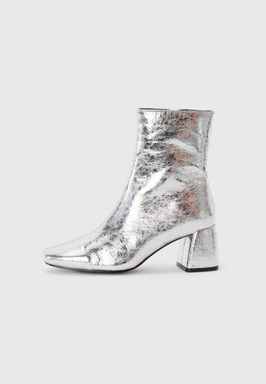 Bottines argentées en matériau brillant et texturé, avec un bout pointu et un design de talon carré. Surface réfléchissante et forme épurée.