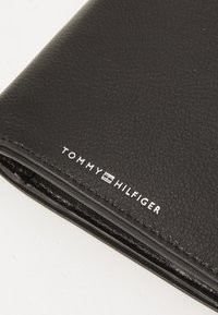 Black leather wallet with a textured finish, featuring white "TOMMY HILFIGER" branding. Zippered closure and stitched edges visible.