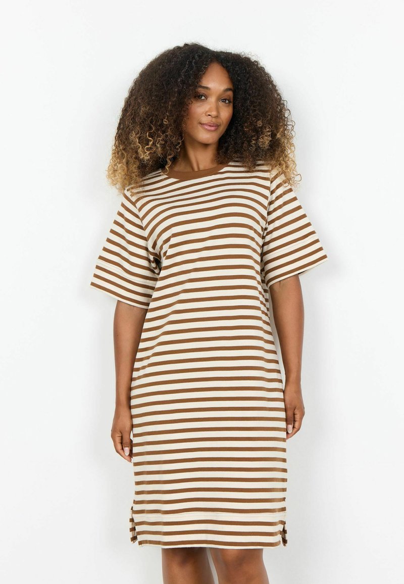Femme aux cheveux bouclés portant une robe à manches courtes et à rayures horizontales marron et blanches jusqu'aux genoux, se tenant devant un fond blanc.