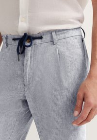 Gestreifte Hosen aus hellblauem und weißem Stoff mit einem Kordelzugbund. Verfügt über seitliche Taschen und eine glatte Textur. Abgerundet mit einem Holzverschluss.