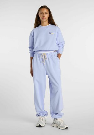 Vrouw staand, gekleed in een lichtpaarse trui en bijpassende joggingbroek met witte sneakers, tegen een effen achtergrond.