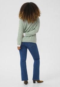 Zielony sweter z prążkowanego materiału z białymi wykończeniami, w połączeniu z niebieskimi spódnicami dzwonami. Figura stoi w brązowych botkach, odwrócona plecami do aparatu.