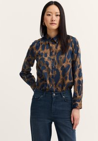Woman with long dark hair wearing gold hoop earrings, a brown and navy patterned button-up shirt, and dark blue jeans against a plain background.