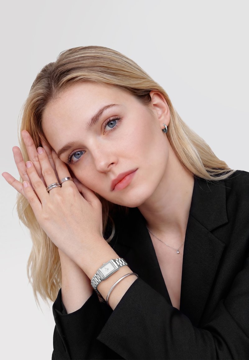 Młoda kobieta o blond włosach, ubrana w czarną kurtkę, opierająca twarz na splecionych dłoniach, nosząca srebrny zegarek, pierścionki, bransoletkę i małe kolczyki.