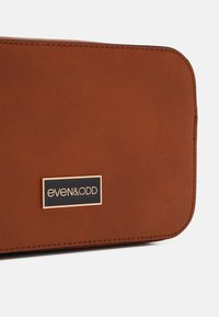 Borsa in finta pelle marrone con una texture liscia, caratterizzata da una forma rettangolare e un logo del marchio accentato in oro su uno sfondo nero.
