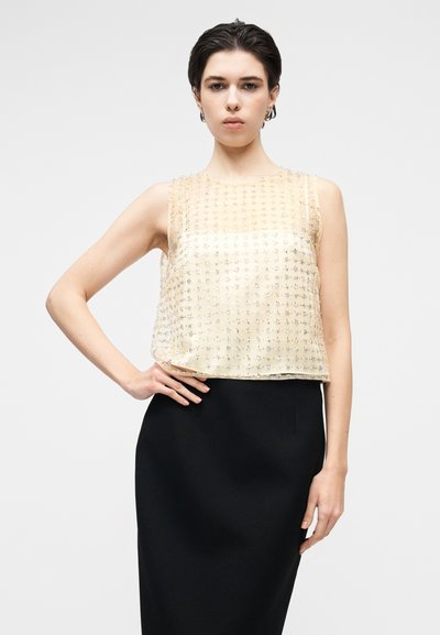 Top bez rękawów w jasnym żółtym kolorze, zdobiony koralikami, z przezroczystym wzorem i metalicznymi akcentami. Połączony z dopasowaną czarną spódnicą.