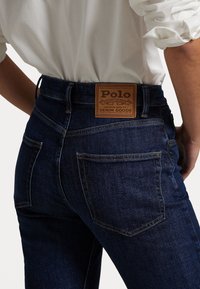 Tmav ě modré džíny s upnutým střihem. Mezi vlastnosti patří hnědá kožená nášivka s logem "Polo" a dvě zadní kapsy.