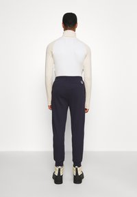 Man som står vänd bort, iklädd en ljusbeige långärmad tröja, mörkblå joggarbyxor och svarta och beige stövlar på vit bakgrund.