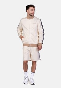 Lichtbeige ritsjack met geribde kraag en boorden, voorzien van zwarte zijstrepen. Gecombineerd met bijpassende beige shorts. Witte sneakers als detail.