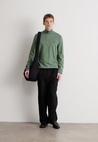 Long-sleeve, light green pullover with a zip neck, paired with black, loose-fitting trousers and a black shoulder bag, worn with black shoes.